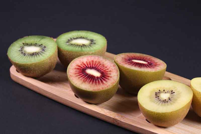 Three varieties of sliced kiwi fruit on a wooden tray on black background