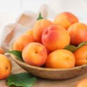 Ripe fresh apricots with green leaves arranged in a wooden bowl on rustic wooden cutting board. Healthy seasonal summer fruit, healthy food, natural nutrition, ingredient for cooking and desserts.
