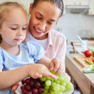 Smiling woman with blond daughter eating fresh grapes in kitchen at home