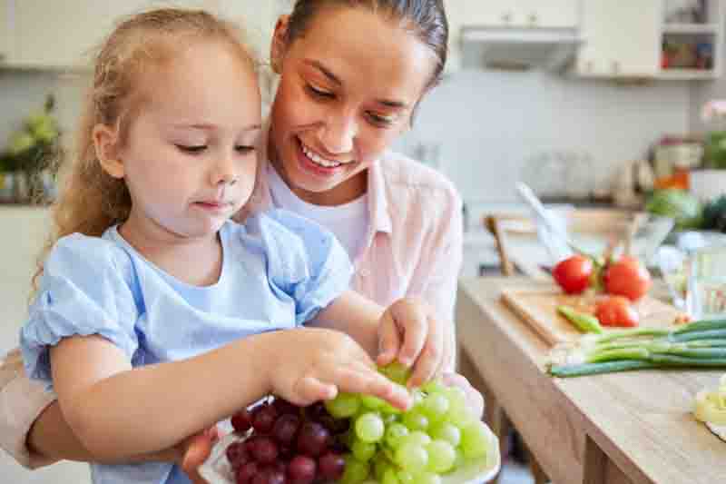 Smiling woman with blond daughter eating fresh grapes in kitchen at home